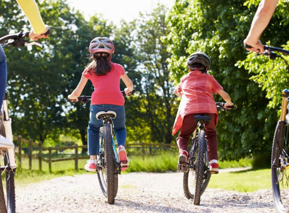 Two cyclists, a boy and a girl riding on a mountain bike, wearing helmets through the lush greenery of Sandwell Valley Country Park, surrounded by trees and other vegetation.