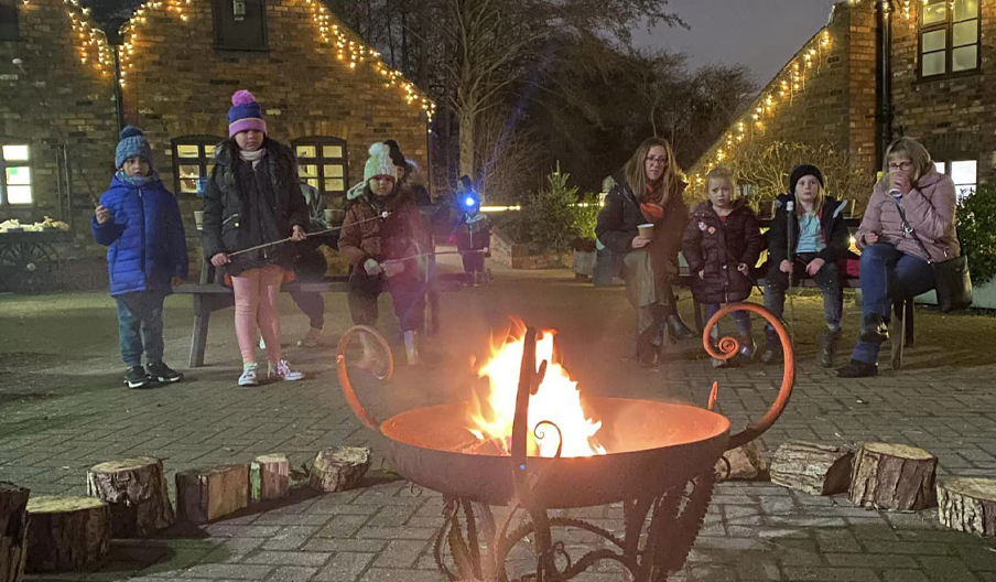 A group of people in winter clothes around a fireplace in the garden where buildings are decorated with Christmas lights.