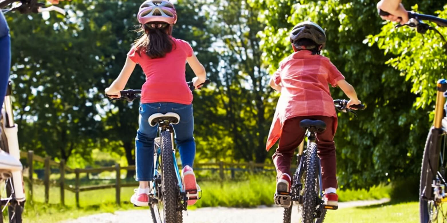 two cyclists, a boy and a girl riding on a mountain bike, wearing helmets through the lush greenery of Sandwell Valley Country Park, surrounded by trees and other vegetation.